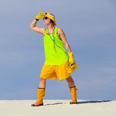 The Atomic Tourist stands facing left, looking out, dressed in assorted neon yellows, holding a Geiger counter