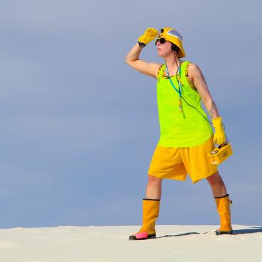 The Atomic Tourist stands facing left, looking out, dressed in assorted neon yellows, holding a Geiger counter