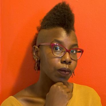 Miatta Kawinzi, a thirty-something Black woman artist, rests her chin on one hand while looking intently at the camera.