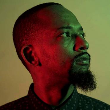 Modesto Flako Jimenez, a thirty-something Afro-Latino with short hair and a beard looking away from the camera while a red glow is on the right side of his face. He is wearing a blue dress shirt with a small pattern.