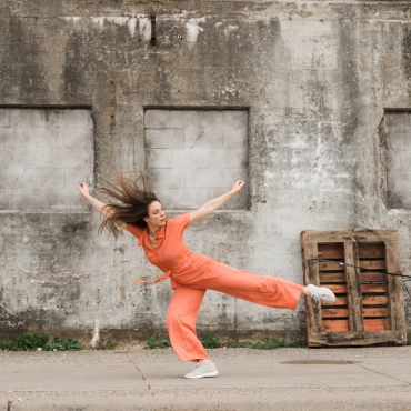 A woman is dancing on a concrete sidewalk near a concrete wall that are white, beige, and black. There are some small green weeds that grow in the the crack in the sidewalk. The woman is dressed in a coral colored jumpsuit that has a collar and short sleeves. She has white skin, brown hair that is blowing in the breeze, and is wearing white tennis shoes. She has one leg extended long towards the back, with all her weight on the other leg. It looks like she is going to take a step back.