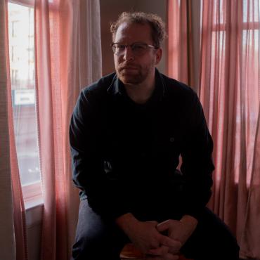 Portrait of a seated man in black shirt with hands clasped in front of pink curtains