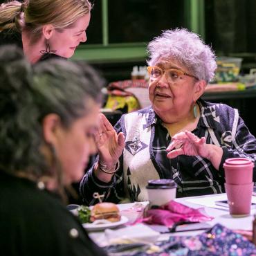 Muriel Miguel at a table, speaking to another woman.