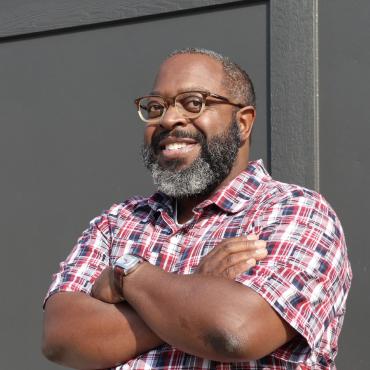 A photo of a middle-aged Black man with grey and white hair, a bushy grey and white beard, and a patterned red, white and blue shirt.