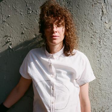 Ka Baird,a 40 something white nonbinary artist with curly brown hair, wearing a white shirt with a circle drawn on it, leans against a textured underpass on a sunny day.