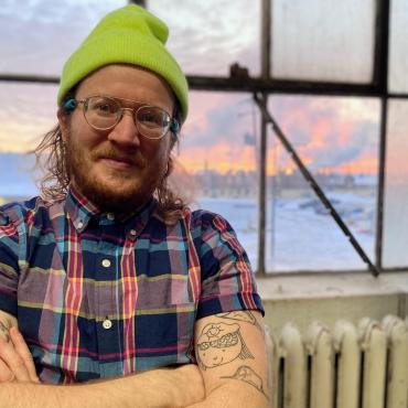  A.P. Looze, a white 30-something transgender person in their studio smiling at the camera.