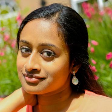 Shruthi Rajasekar, a twenty-something Indian woman, smiling slightly in a sunny field of flowers.