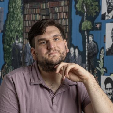 Brooks Turner, a 32 year old white man sits in front of his tapestry "Pantheon" with his head tilted back and fingers at his chin.