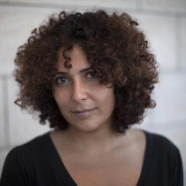 Amber Fares, a Middle Eastern woman with dark curly hair standing in front of a light brick wall.