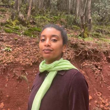 Hasabie, an image of the artist wearing a light green scarf and dark purple sweater. She is taking a walk in the forest with Eucalyptus trees in the background.