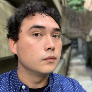 Headshot of the artist with short dark hair wearing a blue shirt. Out of focus in the background is a row of concentric concrete archways and support beams.