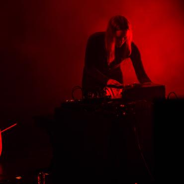 Leila Bordreuil, a 30 year old white woman with medium-long brown hair is facing the camera and playing electronic instruments on a black table. To her right, there is a cello on the floor. She is on a stage with red LED lighting and fog. The main colors of the photo are blacks and reds. 