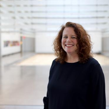 A portrait of Sally, a fair skinned woman with shoulder length curly auburn hair and blue eyes. She is smiling and wearing a black dress with a round neckline. She is standing in the Queens Museum Galleries.