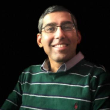 Medium close up photo of a smiling 40 year old Goan Indian man wearing a green and white stripped collared sweater and glasses.