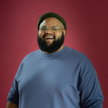 Black man wearing glasses smiles at the camera in front of a red background.