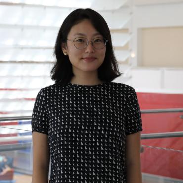 A smiling Korean American woman with short hair, Sarah Cho, stands in the Queens Museum. She wears a black dress patterned with white triangles.