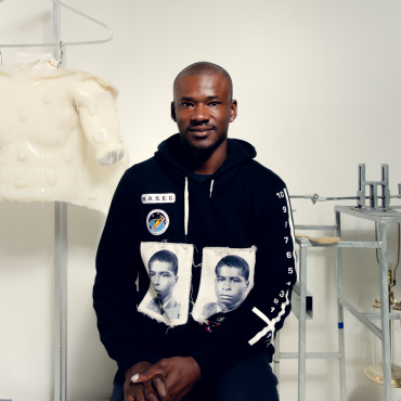 Jeffrey Meris, a mid-30s Afro-Caribbean dark skinned cis-gendered man sits on a stool in his white walled studio. He wears a Black hoodie with white patches designed by artist Tavares Strachan. To his left is a white plaster sculpture coated in silicone that hangs from a metal display rack; to his right is a kinetic sculpture that sits on a table with a head operated by a motor. Three lamps hang below the table.