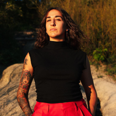 Noelle Awadallah, a young Palestinian woman stoically gazes at the camera with the sun on her skin and the land of Mni Sota Makoce behind her.