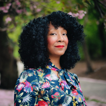 S. Erin Batiste, a 40-something Black woman, red lipped with black curly hair, wearing a blue and watercolor floral print dress, and standing in front of a flowering cherry blossom tree and its overflowing petals.