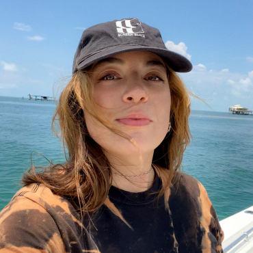 A 30-something filmmaker with long brown hair smiles at the camera. They are wearing a black baseball hat. They are outside on a sunny day and the blue ocean and sky is in the background.