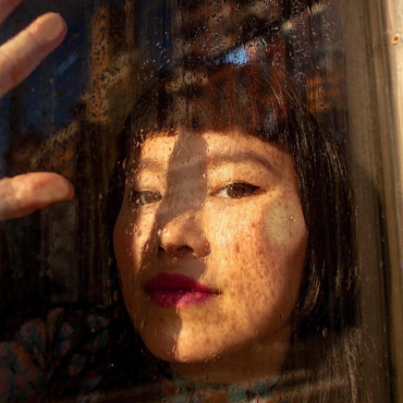 Charmaine Lee, a 30-something Asian woman looking at the camera behind a glass window that has water dripping down it.