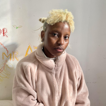 KÌÍTÁN, a Black artist with blonde hair sits at the edge of his bed with Yorùbá inscriptions on the wall behind him.