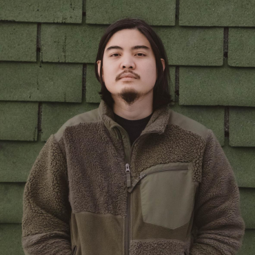 Kengchakaj, a 30-something Thai person with short bob hair and a mustache, wearing a green jacket, standing in front of a green brick-like texture background. He stands and looks directly into the camera with his hand in his pocket.