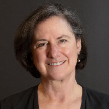 Amy Usdin, a 60-something white woman smiling at the camera in front of a dark grey wall.