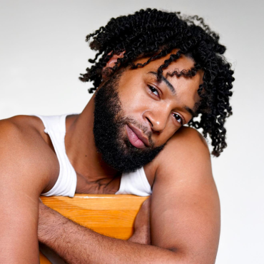 Wendell, a 31-year-old Black male choreographer, hugging a chair and leaning on his left shoulder with a subtle smirk.