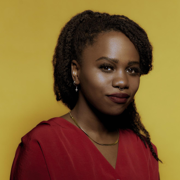 Zola Dee, a late 20's woman, smiles in a red shirt. Behind her is a yellow backdrop.