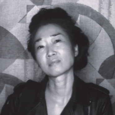 Coleen Baik, a Korean American woman artist wearing a leather jacket, gazes into the camera with a light smile; her head tilted, dark hair in an updo.