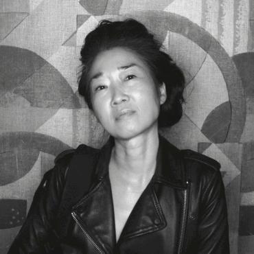 Coleen Baik, a Korean American woman artist wearing a leather jacket, gazes into the camera with a light smile; her head tilted, dark hair in an updo.