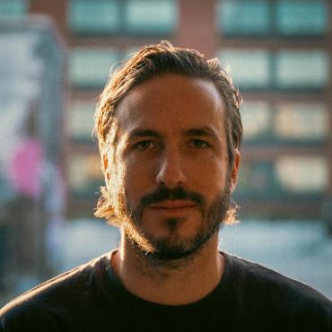 A middle-aged Hispanic man with light skin, giving a slight smile to the camera against an urban background.