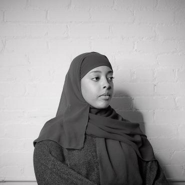 Sara A. Osman, a thirty-year old Somali woman sitting in front of a white wall, wearing hijab and facing slightly away from the camera.