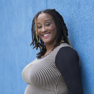 Kayla, a Black voluptuous person, stands with their back against an azure blue concrete wall, and turns their head over their left shoulder to smile joyfully at the camera. They're wearing pink lipstick and their eyebrows are raised in pleasure. Kayla wears a whimsical black and tan thinly-striped shirt.