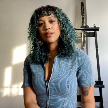Leslie Barlow, a 36-year-old Black woman with light brown skin, two facial piercings, and curly teal-colored hair smiling in front of the white wall in her studio, with a painting easel just behind her and to the right.