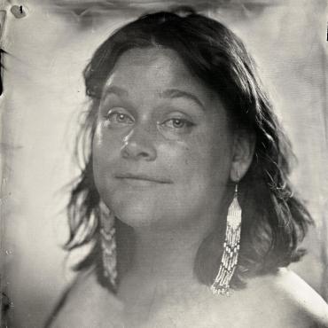A black and white tintype portrait of Halee Kirkwood, a thirty-something mixed white and Ojibwe poet with shoulder-length hair and long beaded earrings. They wear a black t-shirt while slightly smiling at the camera.