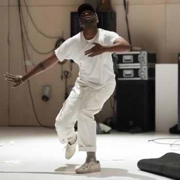 Jordan Demetrius Lloyd, A 31-year old dancer dressed in white, looks up with bent knees and extended arms.