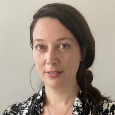 Headshot of the artist with dark hair tied back, looking at the camera, wearing a black-and-white patterned button-up shirt.